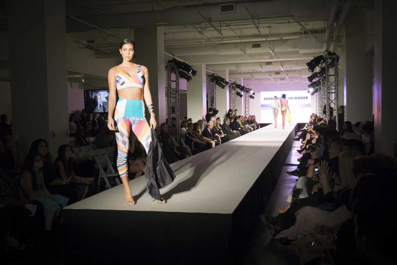 Model walking a long white runway in a dark hall lined with guests