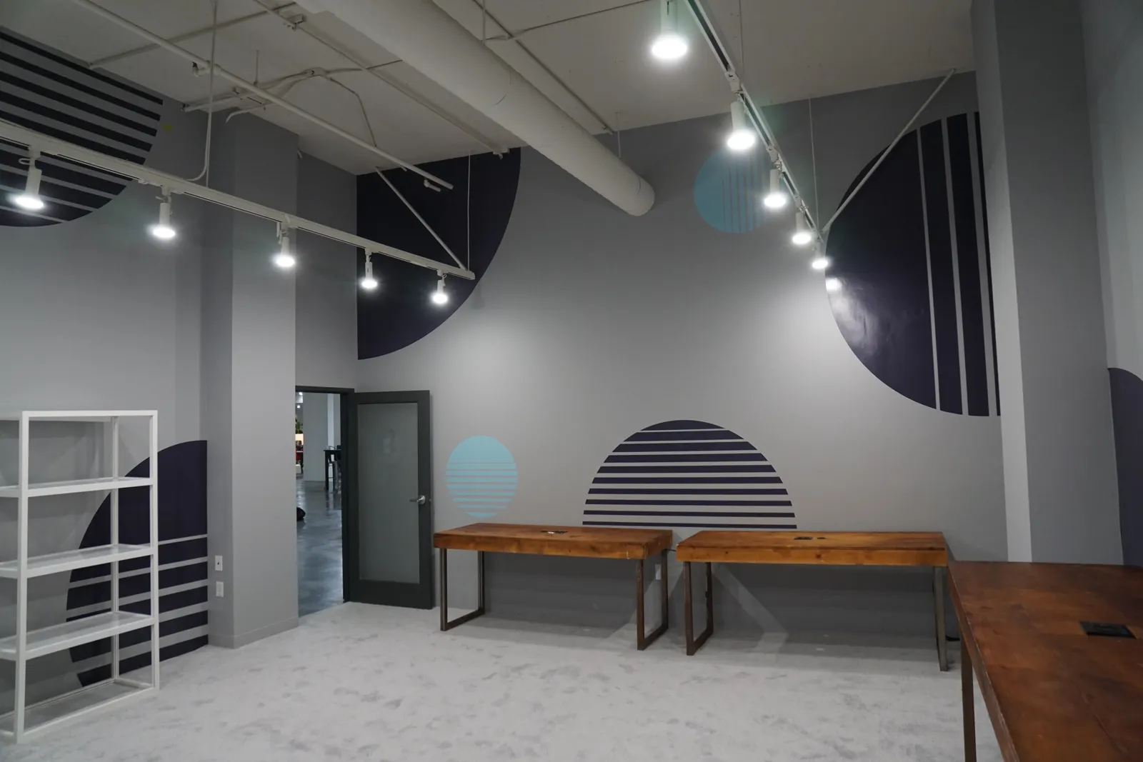 Plain grey room with track lights, wooden tables and an open shelf