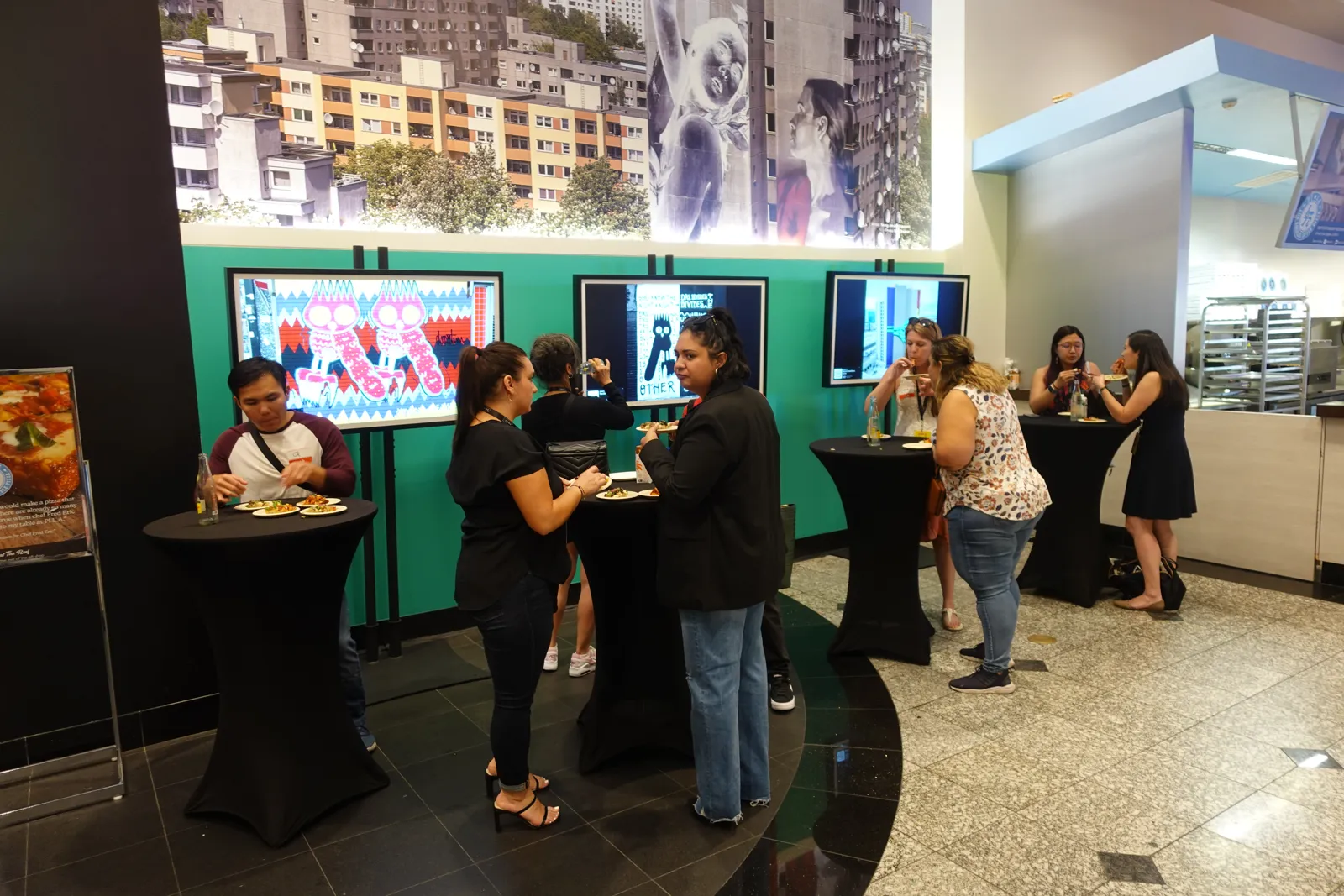 Guests with drinks at cocktail tables in a lobby with green walls, screens and a city mural