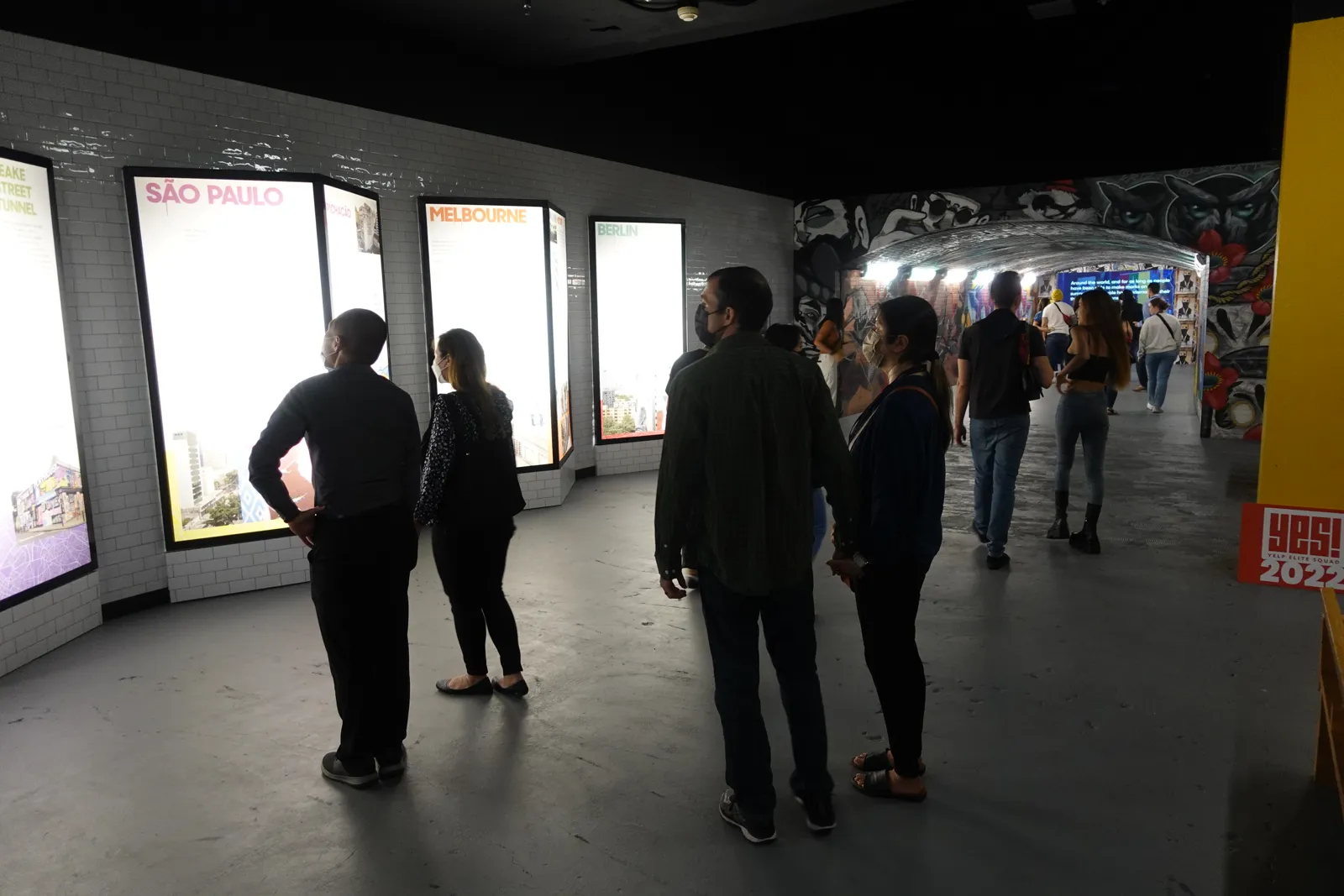Visitors stand by lit city scene lightboxes in a corridor leading to a graffiti tunnel