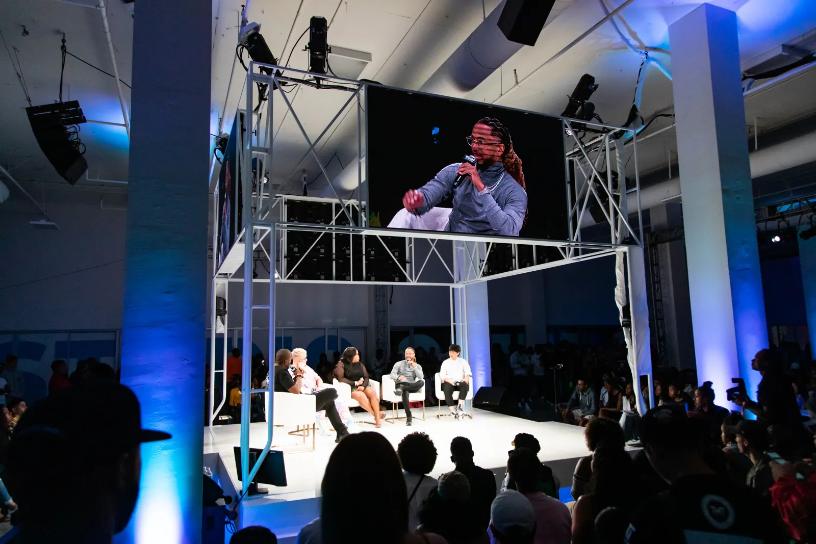 Panel of speakers seated on a white stage beneath a large video screen