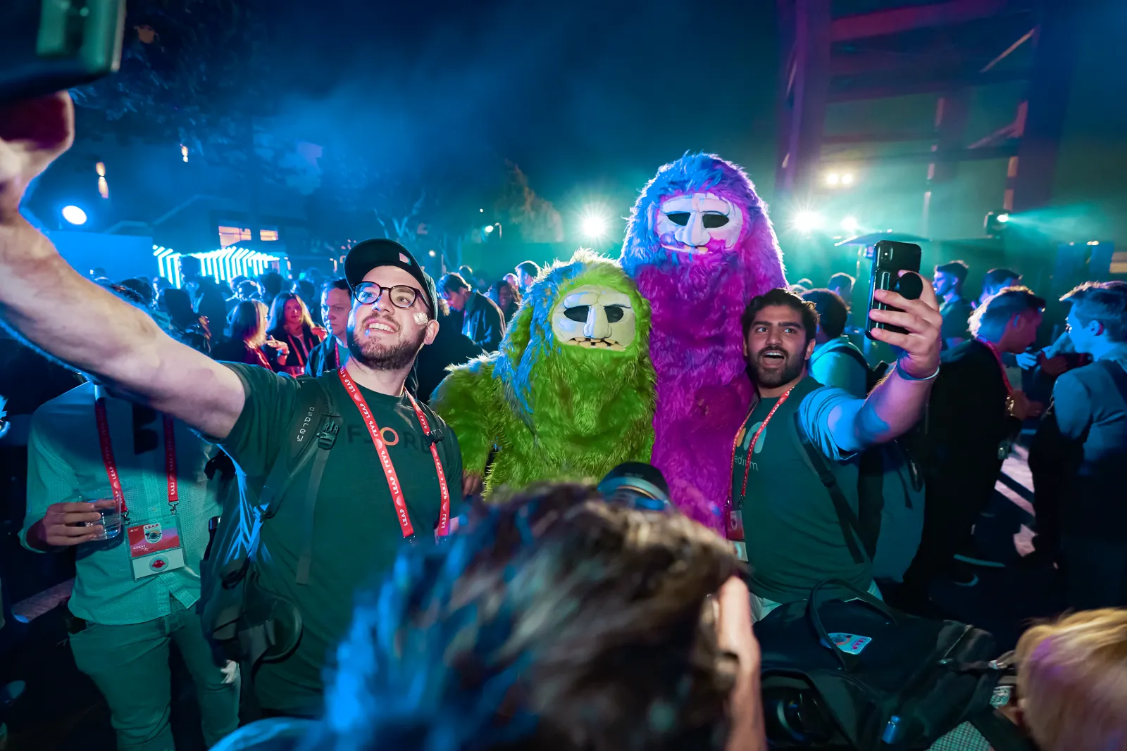 Crowd taking selfies with costumed mascots under green stage lights