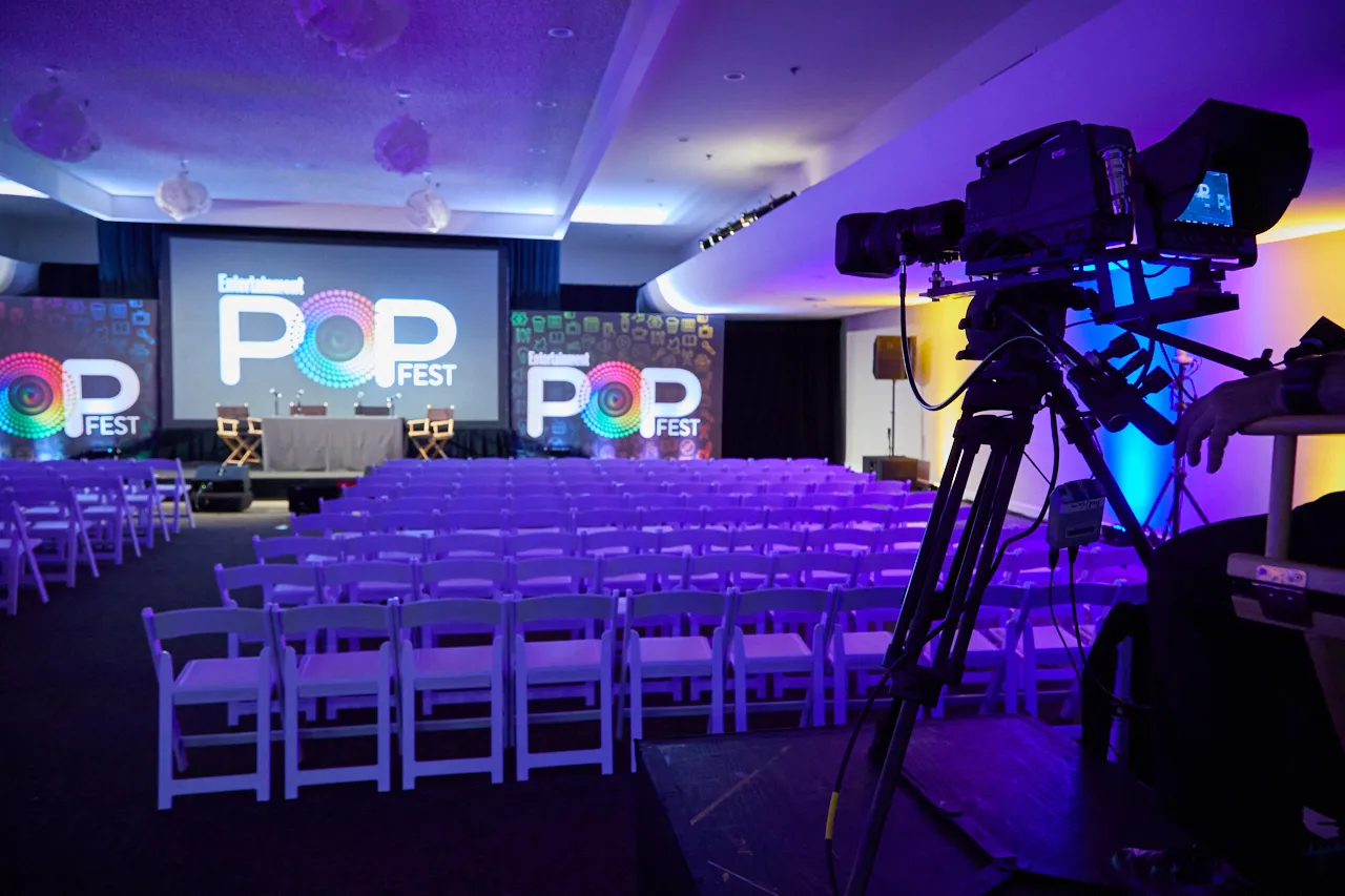 Rows of white folding chairs facing a stage screen, with a camera in front