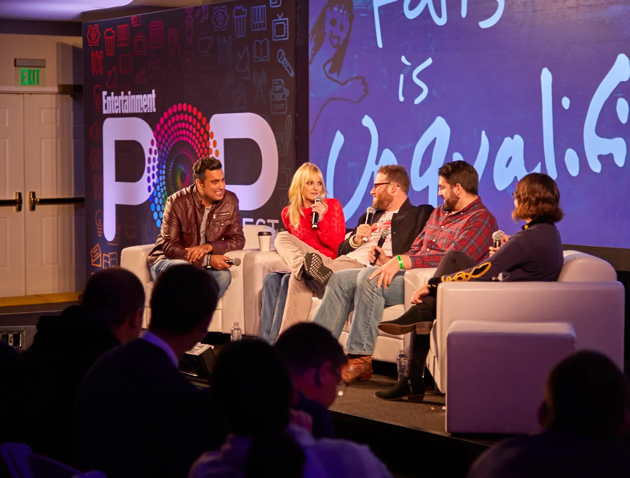 Five panelists on white sofas speaking to an audience from a lit stage