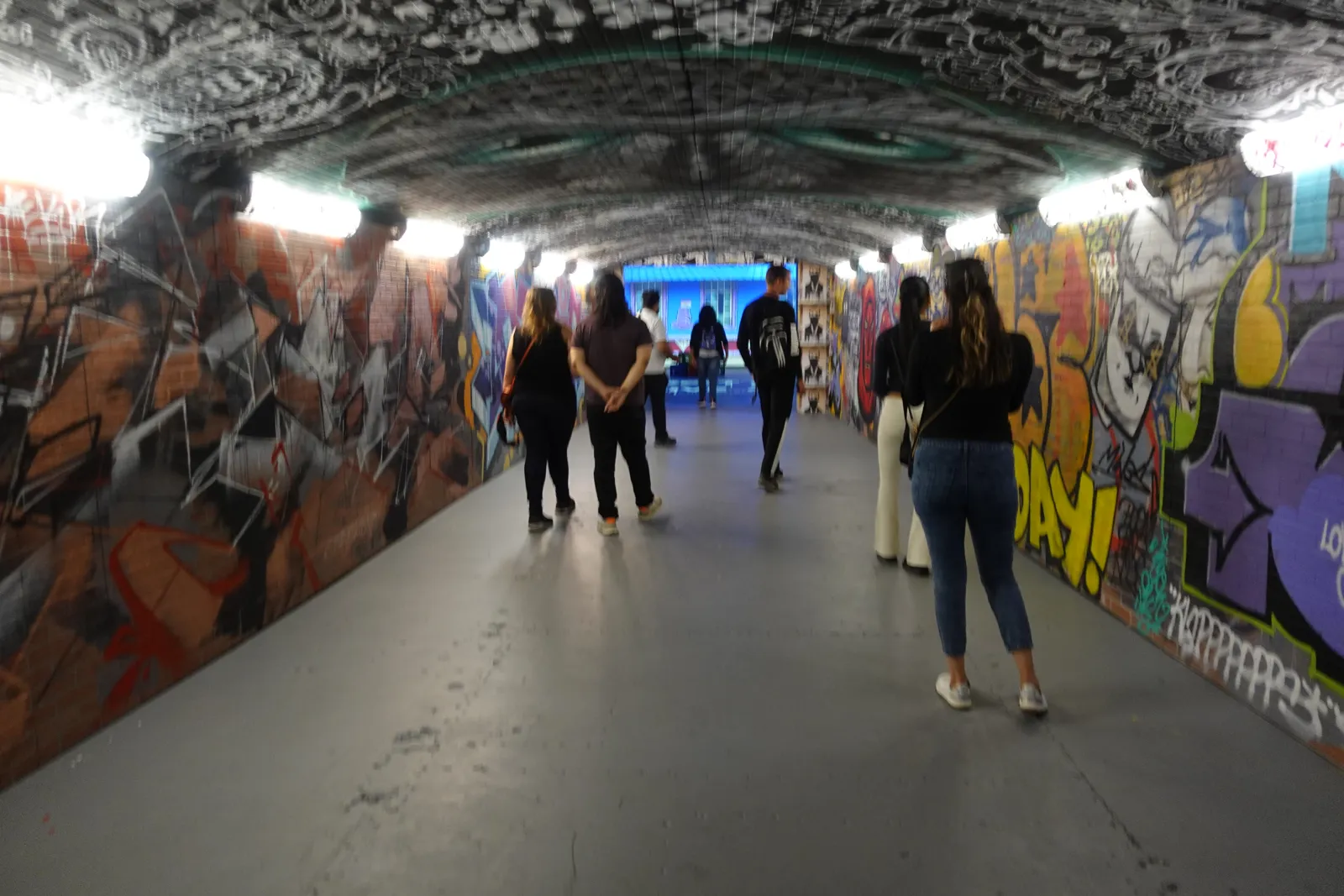 People walking through a graffiti covered tunnel under bright overhead lights