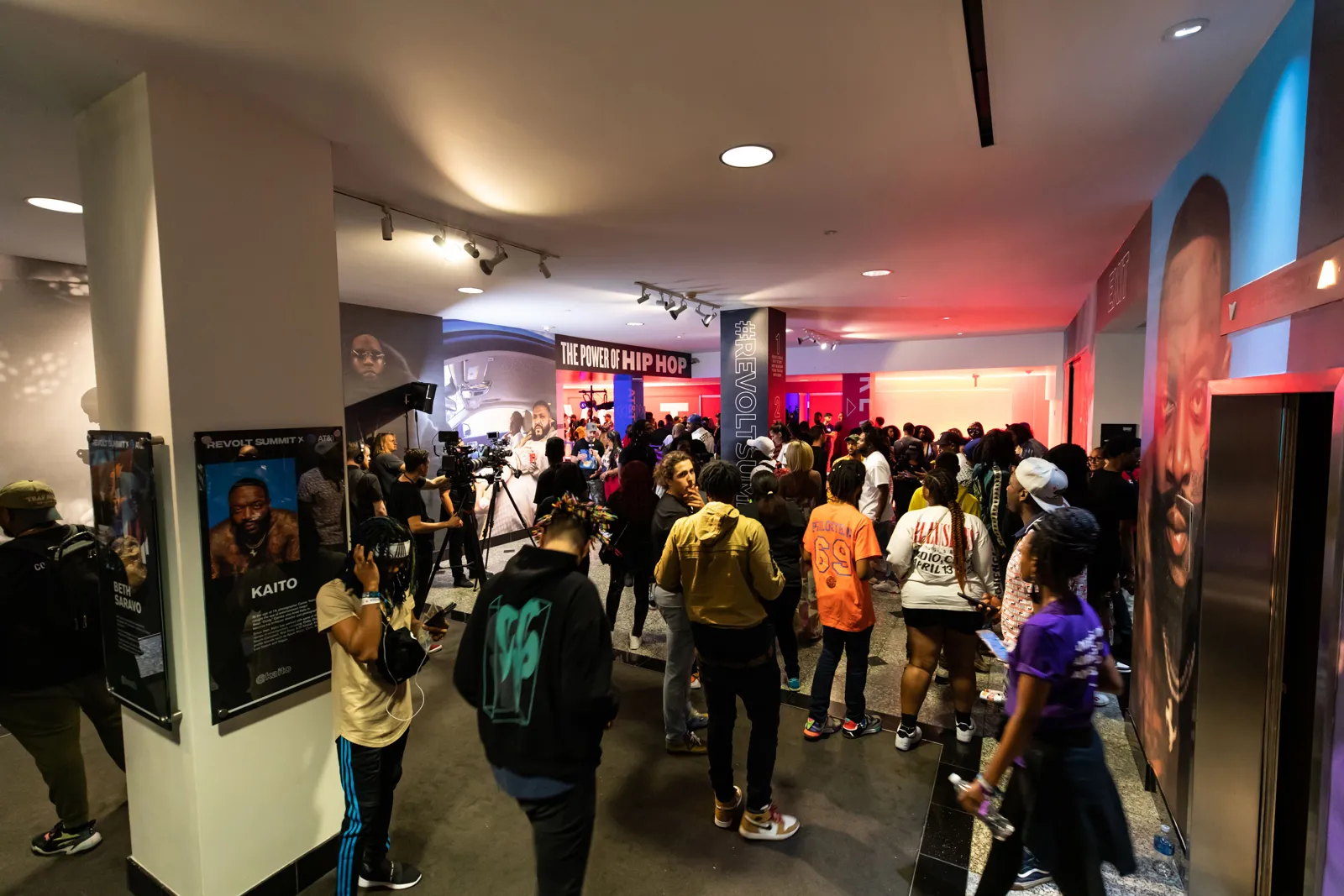 Busy crowd moving through a gallery hallway lined with portraits and lights