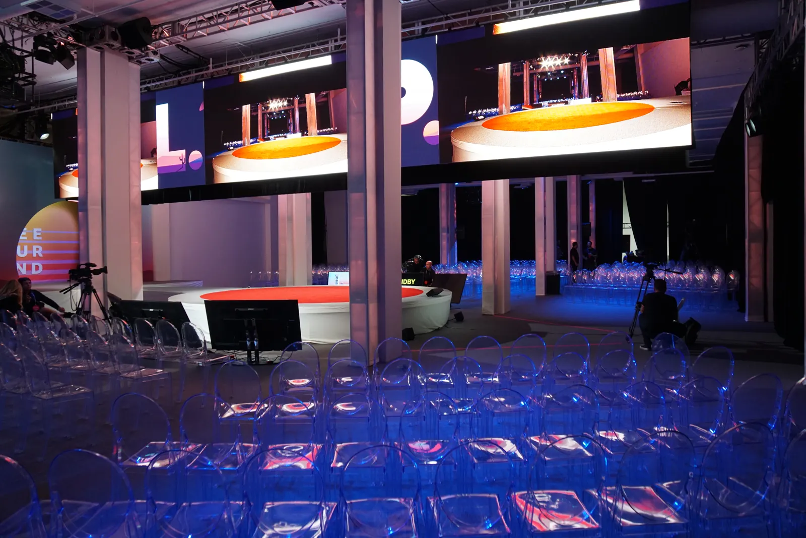 Rows of clear chairs facing a round stage beneath large video screens